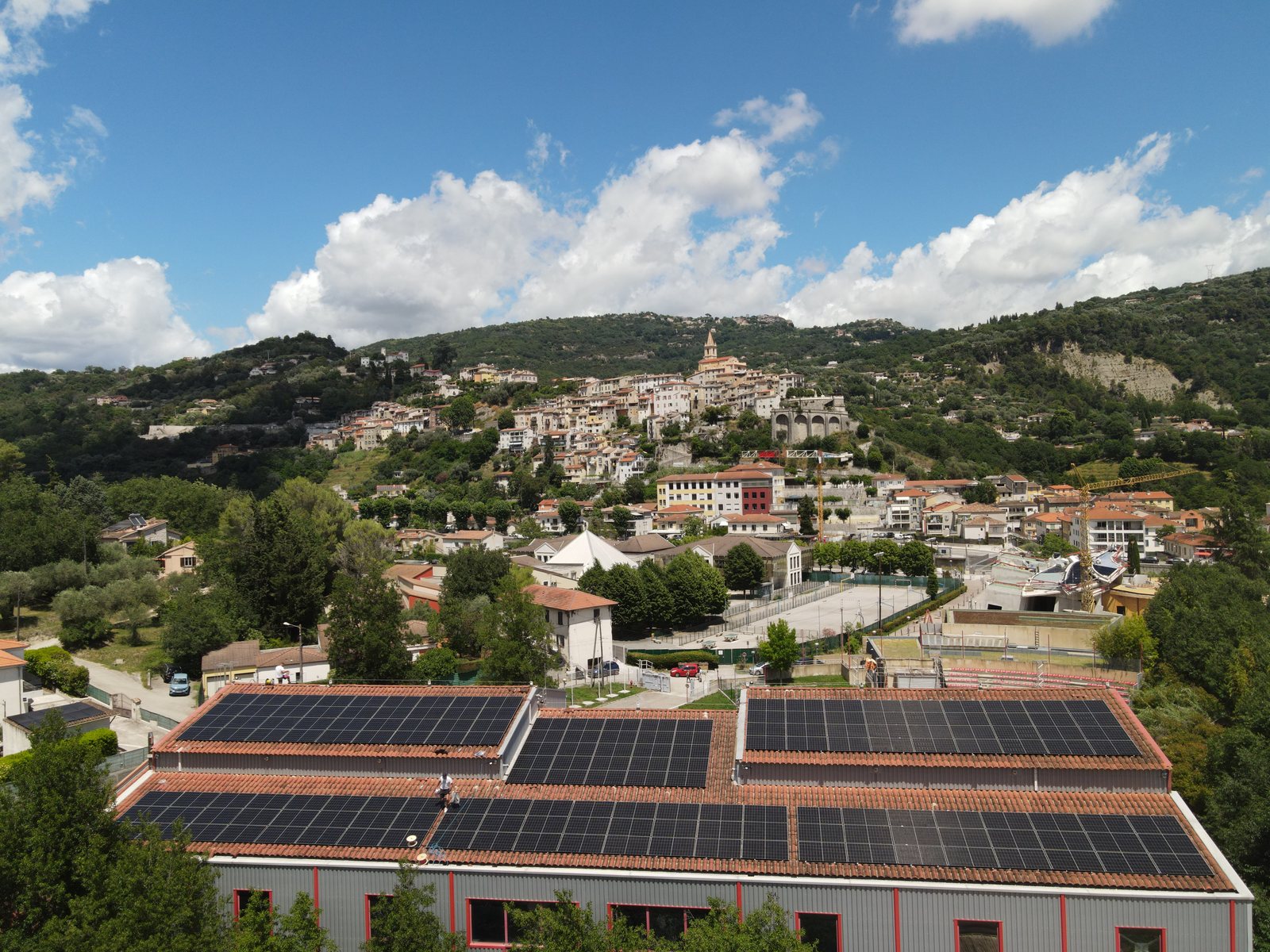 Gymnase municipal de Contes équipé de modules photovoltaïques, avec le village perché en arrière-plan, vue de drone